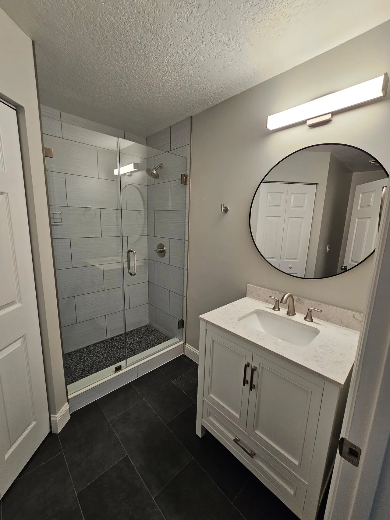 Finished shower with gray plank tile, a river-rock floor and frameless glass, next to a white vanity